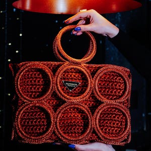 May include: A handmade, rust-colored crochet handbag with a circular handle and decorative circular accents. The bag features a small, triangular tag that reads "HANDMADE Fischer.co". The bag is held up by a person with blue nail polish.