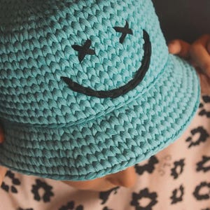 Peut inclure: Un chapeau bob en crochet bleu clair orn&eacute; d'un visage souriant noir brod&eacute; avec deux yeux en forme de X et une bouche incurv&eacute;e. Le chapeau est fabriqu&eacute; &agrave; partir d'un fil &eacute;pais et textur&eacute; avec une calotte arrondie et un bord inclin&eacute;.