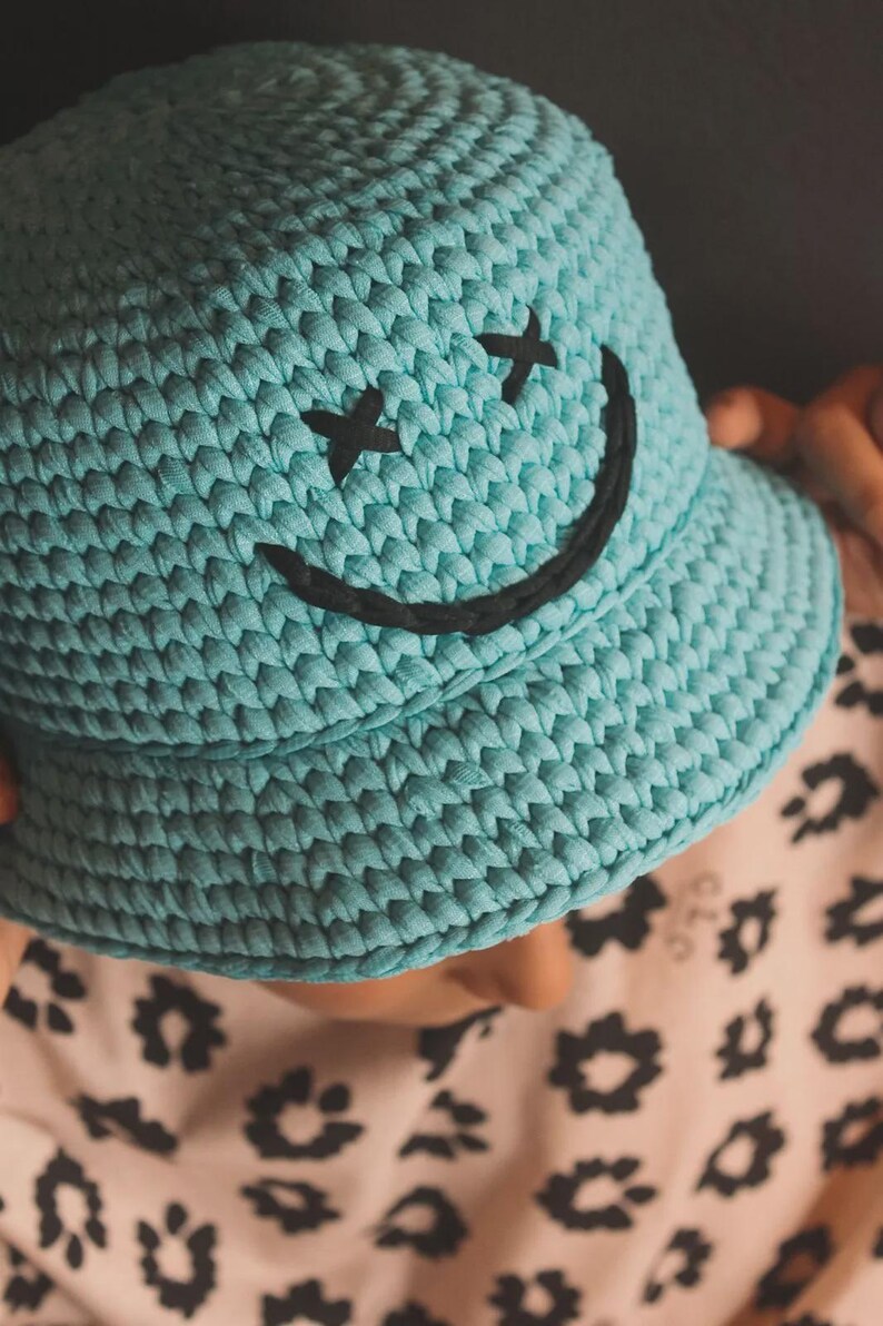 Peut inclure: Un chapeau bob en crochet bleu clair orn&eacute; d'un visage souriant noir brod&eacute; avec deux yeux en forme de X et une bouche incurv&eacute;e. Le chapeau est fabriqu&eacute; &agrave; partir d'un fil &eacute;pais et textur&eacute; avec une calotte arrondie et un bord inclin&eacute;.
