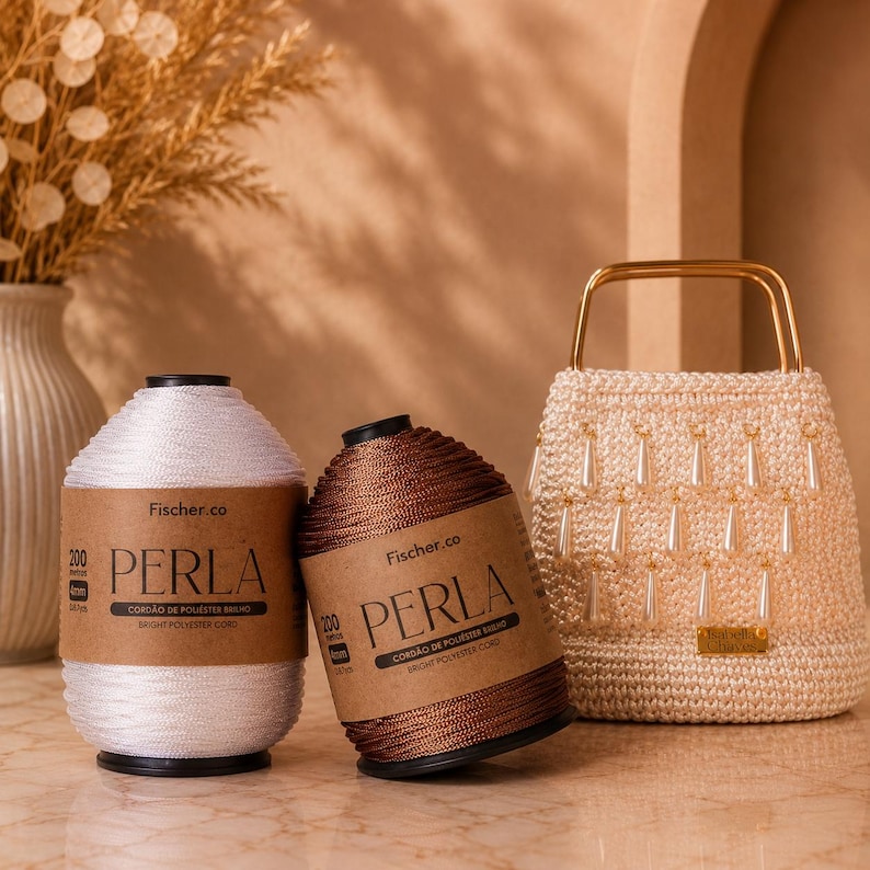 May include: Two spools of Fischer.co Perla bright polyester cord, one white and one bronze, each labeled 200 meters and 4mm, positioned next to a cream-colored crocheted handbag featuring gold-tone metal handles and dangling white teardrop beads.
