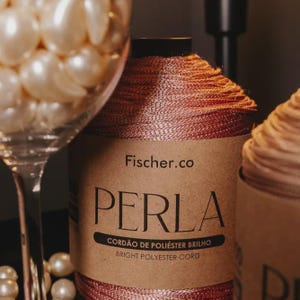 May include: A spool of metallic rose-gold polyester cord with a paper label reading Fischer.co PERLA CORD&Atilde;O DE POLI&Eacute;STER BRILHO BRIGHT POLYESTER CORD, positioned next to a clear glass filled with numerous round, cream-colored faux pearls.