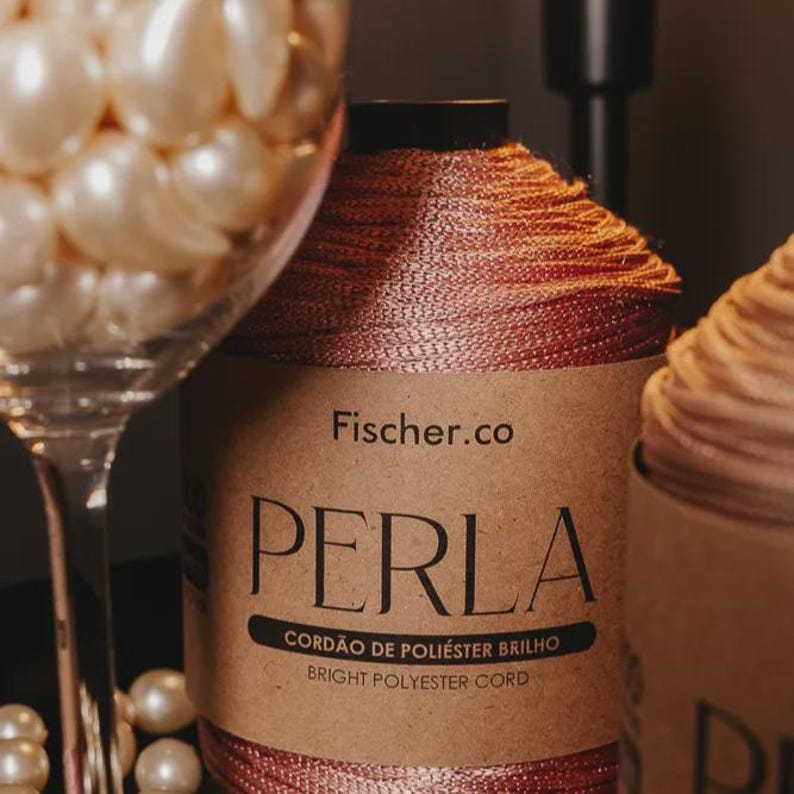 May include: A spool of metallic rose-gold polyester cord with a paper label reading Fischer.co PERLA CORD&Atilde;O DE POLI&Eacute;STER BRILHO BRIGHT POLYESTER CORD, positioned next to a clear glass filled with numerous round, cream-colored faux pearls.