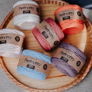 Peut inclure: Six bobines de fil Refinatto en blanc, rose clair, bleu clair, rose fonc&eacute;, orange et violet. Chaque bobine porte une &eacute;tiquette en papier brun avec le texte Refinatto, 140m et 15mm. Le fil est dispos&eacute; en cercle sur un plateau rond.
