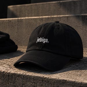 Könnte beinhalten: Eine schwarze Baseballkappe mit gebogenem Schirm und strukturierter Krone. Auf der Vorderseite ist das Wort "jebiga." in weißen Kleinbuchstaben eingestickt. Die Kappe hat einen Knopf in der Mitte oben und sichtbare Nähte am Schirm.