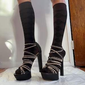 Op de afbeelding: Zwarte plateau sandalen met dikke gevlochten banden en een chunky hak. De sandalen worden gedragen met zwarte en witte ruitvormige kniekousen.