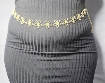 May include: A gold chain belt with small, white flower-shaped charms. The belt is worn around the waist of a person wearing a black ribbed dress.