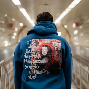 May include: A blue hoodie featuring a graphic print of a woman's face and the text "I always been a top-notch Hollywood Superstar in my mind."