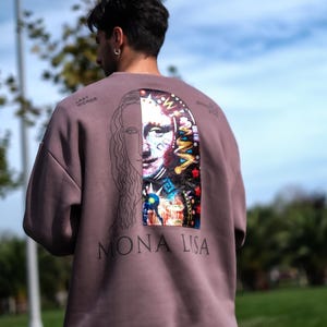 May include: A brown sweatshirt with a black outline of the Mona Lisa on the back. The right side of the image is filled with a colourful, abstract design. The text "Mona Lisa" is printed below the image. The text "Last Words" is printed on the left shoulder of the sweatshirt.
