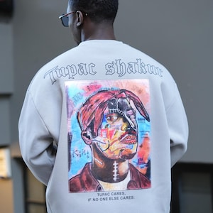 May include: A light gray sweatshirt with a colorful graphic print of Tupac Shakur's face on the back. The text "Tupac Shakur" is printed in black on the back of the sweatshirt. The text "THUG LIFE" is printed in white on the graphic. The text "TUPAC CARES, IF NO ONE ELSE CARES." is printed in black below the graphic.