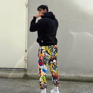K&ouml;nnte beinhalten: Eine Person tr&auml;gt ein schwarzes Kapuzenpullover und eine bunte Hose mit grafischem Muster. Die Hose hat einen wei&szlig;en Hintergrund mit verschiedenen Farben und Designs, darunter Gelb, Rot, Blau und Gr&uuml;n. Die Person tr&auml;gt wei&szlig;e Turnschuhe.