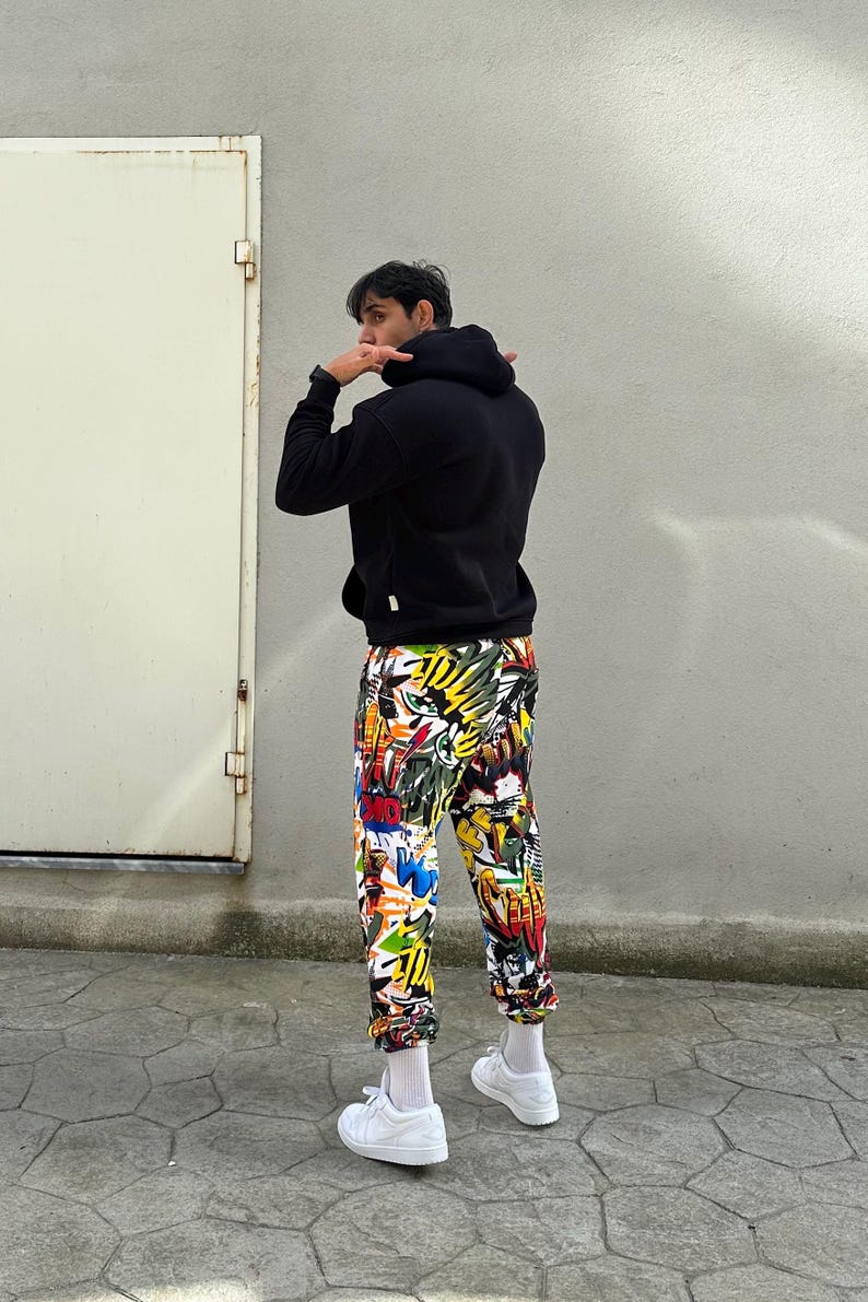K&ouml;nnte beinhalten: Eine Person tr&auml;gt ein schwarzes Kapuzenpullover und eine bunte Hose mit grafischem Muster. Die Hose hat einen wei&szlig;en Hintergrund mit verschiedenen Farben und Designs, darunter Gelb, Rot, Blau und Gr&uuml;n. Die Person tr&auml;gt wei&szlig;e Turnschuhe.