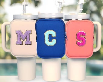 Puede incluir: Tres vasos térmicos con fundas de neopreno. Las fundas son de color crema, azul real y coral. Cada funda tiene una letra grande, M, C y S, en un color contrastante con detalles de purpurina.