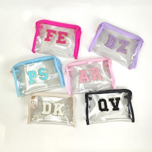 May include: Six clear plastic zippered pouches with different coloured fabric trim. Each pouch has a large, fluffy letter appliqué with gold glitter accents. The letters spell out "FE", "BZ", "PS", "AR", "DK", and "QV".