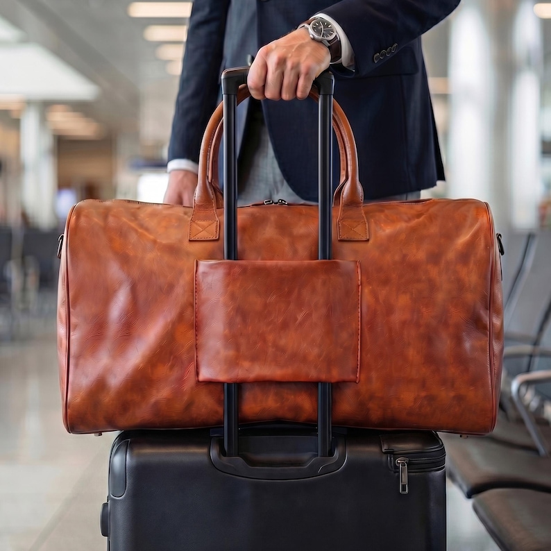 May include: A large, rectangular, cognac-colored leather duffel bag with two top handles and a central luggage sleeve. The bag is positioned horizontally, resting on top of a black rolling suitcase with an extended handle, featuring a smooth finish.