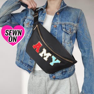 May include: A black fanny pack with a gold zipper and the word "AMY" spelled out in colorful letters. The fanny pack is worn by a person wearing a denim jacket.