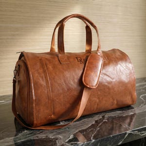 May include: A tan duffel bag with a smooth, textured finish, featuring two top handles, a detachable shoulder strap with a padded cushion, and the letters DM embossed on the front. The bag has a rectangular shape with rounded edges and side zippers.