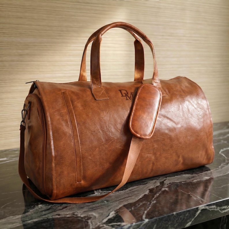 May include: A tan duffel bag with a smooth, textured finish, featuring two top handles, a detachable shoulder strap with a padded cushion, and the letters DM embossed on the front. The bag has a rectangular shape with rounded edges and side zippers.