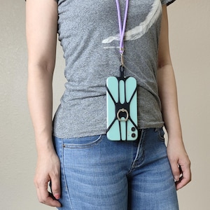 May include: A light mint green smartphone encased in a black silicone harness with an integrated circular metal ring stand. The device is attached to a lavender cord lanyard that hangs around the neck, worn over a gray t-shirt and blue jeans.