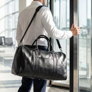 May include: A black leather duffel bag with two top handles and a detachable shoulder strap. The bag features a horizontal front pocket and the name James embossed in silver lettering on the front. The bag has a smooth, polished finish.