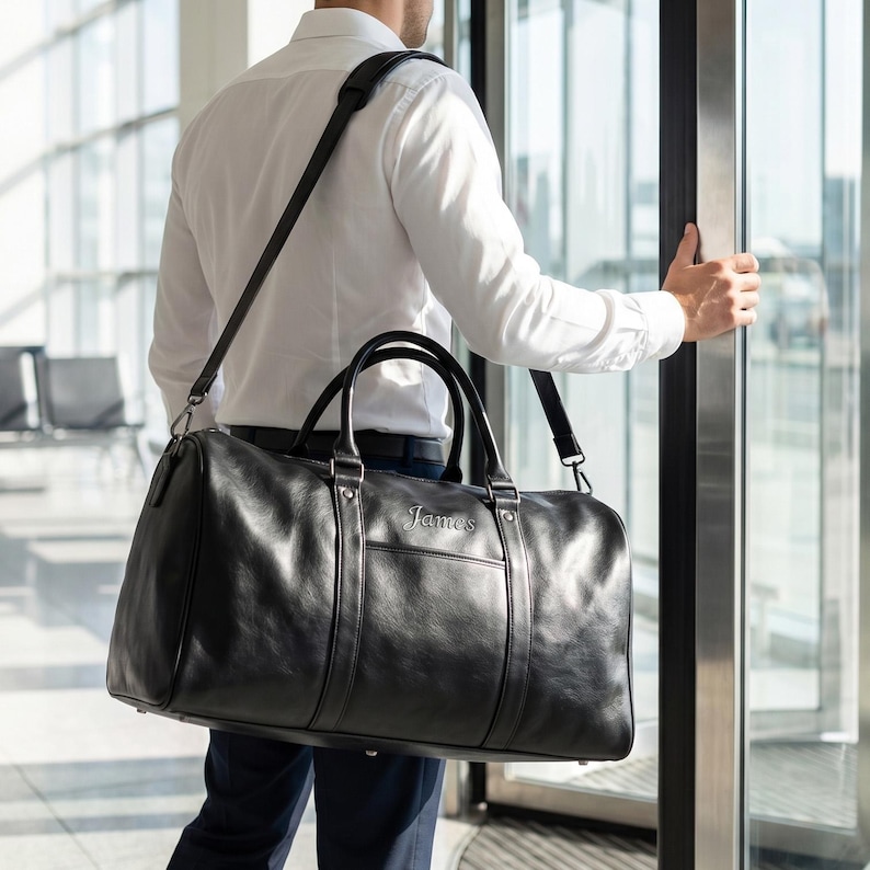 May include: A black leather duffel bag with two top handles and a detachable shoulder strap. The bag features a horizontal front pocket and the name James embossed in silver lettering on the front. The bag has a smooth, polished finish.