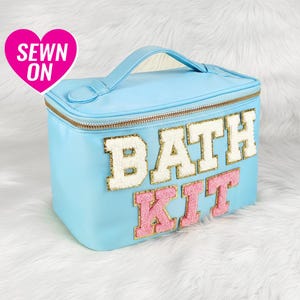 Puede incluir: Un neceser rectangular azul claro con asa y cremallera dorada. Las palabras "BATH KIT" están cosidas en la parte delantera con letras blancas y rosas. Un corazón rosa con las palabras "SEWN ON" está en la esquina superior izquierda.