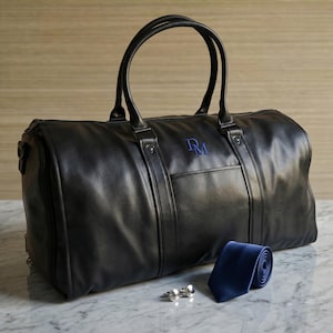 May include: A black duffel bag with two top handles and a blue embroidered monogram reading DM. Beside the bag lies a rolled solid blue necktie and a pair of silver-toned metal cufflinks. The bag features a smooth finish and a front pocket.