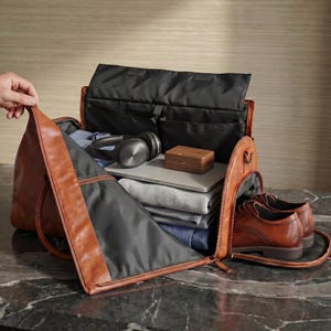 May include: A brown leather duffel bag with a black interior lining, containing folded grey and blue clothing, a silver laptop, black over-ear headphones, a small wooden box, and a pair of brown leather dress shoes in a side compartment.