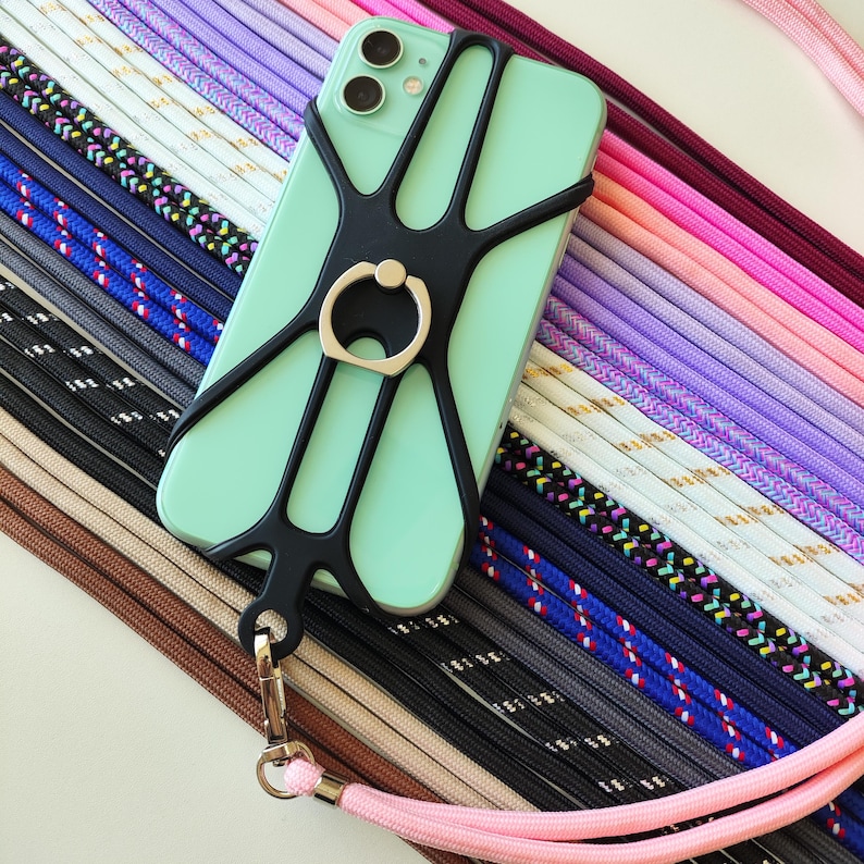 May include: A mint green phone case with a black spider web design and a silver ring holder. The case is attached to a pink lanyard with a black and white striped pattern.