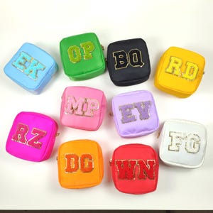 May include: A selection of small, square cosmetic bags in various colours, each decorated with sequined block letters. Colours include blue, green, black, yellow, pink, purple, orange, red, and white. The bags have gold-toned zips.