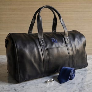 May include: A black leather duffel bag featuring two top handles, a zippered closure, and the blue embroidered monogram letters DM on the front. Beside the bag lies a rolled solid blue necktie and a pair of polished silver-toned metal cufflinks.