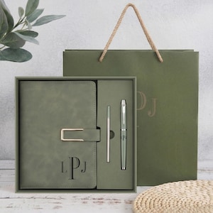 May include: A sage green notebook with a gold-toned rectangular clasp and the monogram LPJ embossed in black, paired with a matching pen in a gift box. The set includes a coordinating sage green paper gift bag with rope handles and the monogram LPJ.