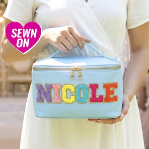Peut inclure: Une trousse de maquillage bleu clair avec une fermeture éclair et une poignée dorées. Le nom "NICOLE" est cousu sur le devant en lettres colorées et texturées. Un autocollant rose en forme de cœur avec les mots "SEWN ON" est en haut à gauche.
