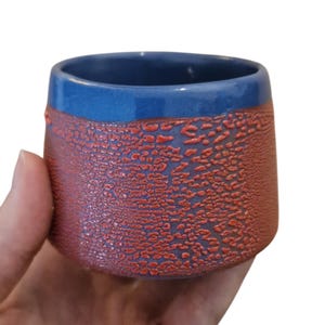 Könnte beinhalten: Ein kleiner Keramikbecher mit einem glatten, glänzenden blauen Rand und einem strukturierten, rotbraunen Unterteil. Die Oberfläche des unteren Bereichs weist ein ausgeprägtes, erhabenes, rissiges Muster auf, das mit dem blauen Band kontrastiert.