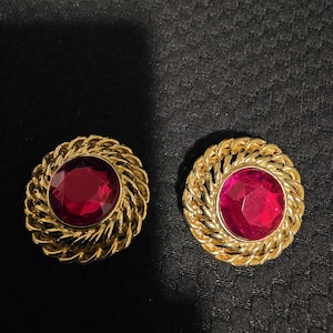May include: A pair of gold-toned clip-on earrings. Each earring features a large, round, ruby-red gemstone set within a twisted rope-like frame. The earrings are displayed on a dark, textured surface.