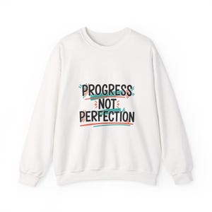 May include: White crew neck sweatshirt with the text "PROGRESS NOT PERFECTION" in black, teal, and red. The sweatshirt is a solid white color and has long sleeves. The text is centered on the front of the sweatshirt.