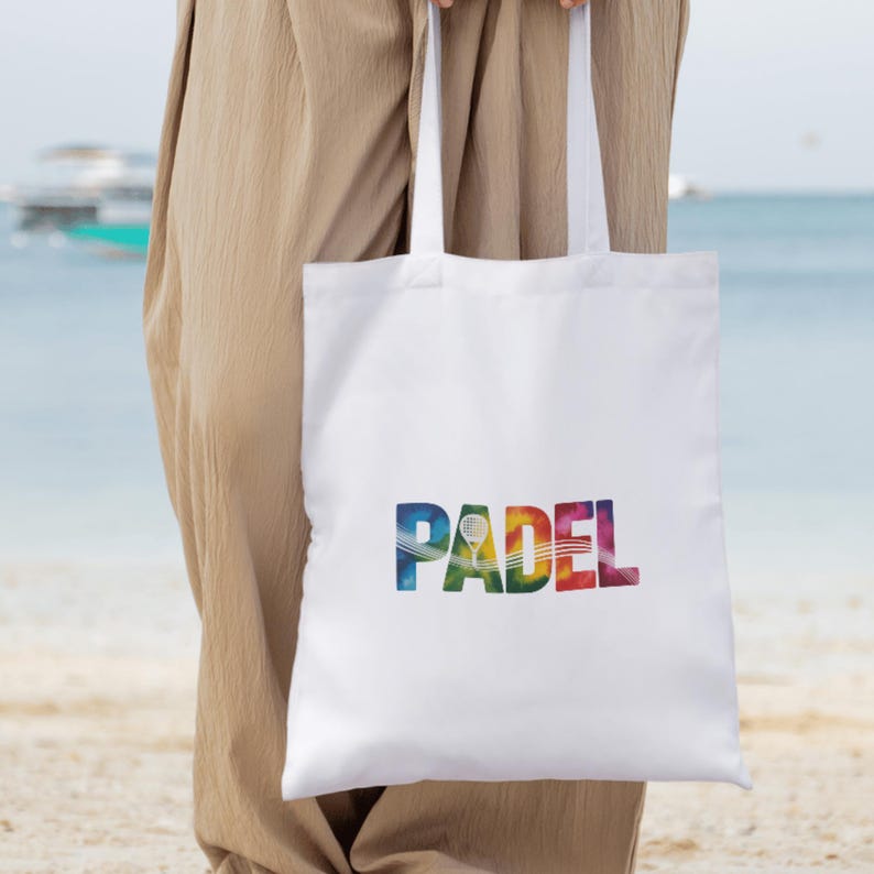 May include: White tote bag with the word "PADEL" in colorful, rainbow-style lettering. A tennis racket graphic is incorporated into the design. The bag has white handles and is held by a person.