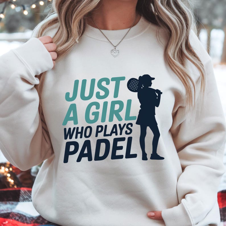 May include: Cream-colored sweatshirt with the text "JUST A GIRL WHO PLAYS PADEL" in teal and navy blue. A silhouette of a person holding a padel racket is on the right side of the text. The person is wearing a hat and skirt.