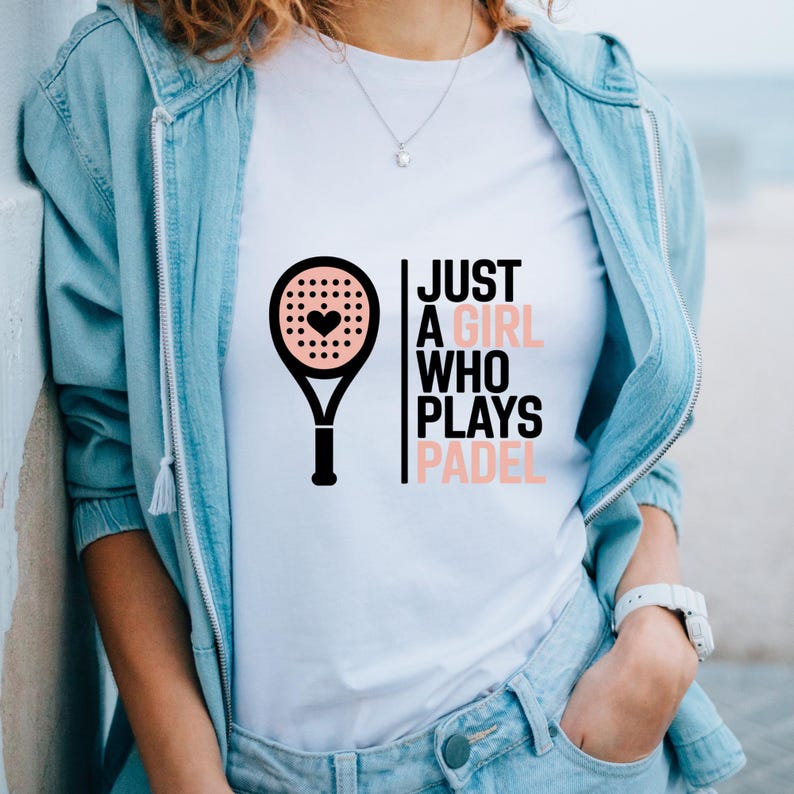 May include: White t-shirt featuring a black and pink padel racket graphic with a heart in the center. The text reads "JUST A GIRL WHO PLAYS PADEL" in black and pink. The shirt is worn with a light blue jacket and jeans.