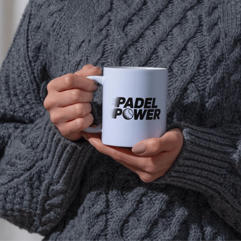 May include: A white ceramic mug with the words "PADEL POWER" printed in black, along with a stylized image of a tennis ball. The mug is held by a person wearing a gray sweater.