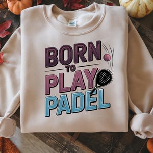 May include: A beige sweatshirt with the text "BORN TO PLAY PADEL" in purple, pink, and blue. A padel racket and ball are illustrated next to the text. The sweatshirt has long sleeves and a crew neck.