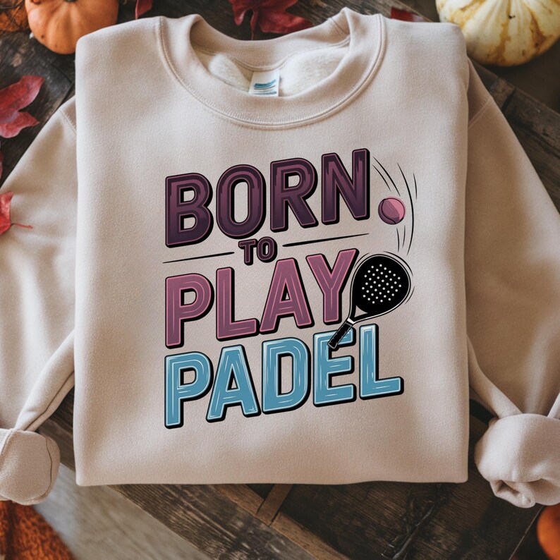 May include: A beige sweatshirt with the text "BORN TO PLAY PADEL" in purple, pink, and blue. A padel racket and ball are illustrated next to the text. The sweatshirt has long sleeves and a crew neck.