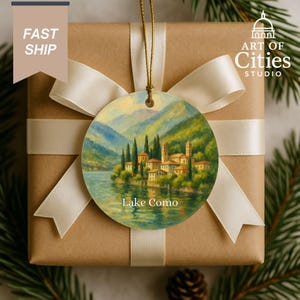 May include: A round Christmas ornament featuring a painting of Lake Como, Italy, with a scenic view of the lake, mountains, and buildings. The ornament is suspended by a gold string and is set against a brown gift box with a white ribbon. The text "Lake Como" is visible.