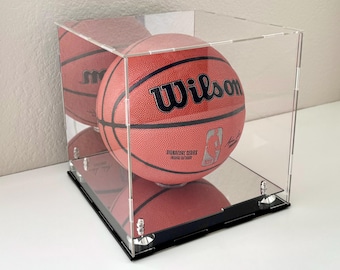 May include: A red Wilson Signature Series basketball inside a clear acrylic display case with a black base. The basketball has black lettering and the NBA logo. The case is square and sits on a white surface.