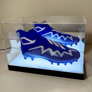May include: A pair of blue and white football cleats displayed in a clear acrylic case with a black base. The cleats have a unique zigzag design and are illuminated by a soft, white light.