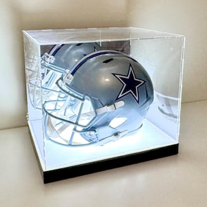 May include: A clear acrylic display case with a black base, showcasing a silver football helmet with a blue star on the side. The helmet is illuminated from below by a white LED light.