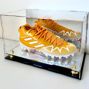 May include: A pair of gold and white football cleats displayed inside a clear acrylic case with a black base. The cleats have white accents and orange laces. The case has a mirrored back, reflecting the cleats.