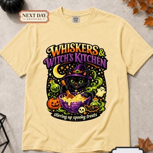 May include: A butter yellow short-sleeve t-shirt featuring a graphic of a black cat wearing a witch hat, stirring a purple bowl. The text reads Whiskers & Witch's Kitchen and stirring up spooky treats, surrounded by a pumpkin, potions, and a ghost.
