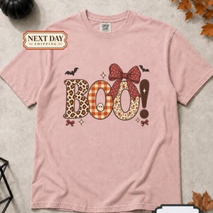 May include: A dusty pink short-sleeve t-shirt featuring the word BOO! in large letters. The B has a leopard print, the O has an orange gingham pattern with a small ghost, and the second O has a floral pattern with a large red polka dot bow.