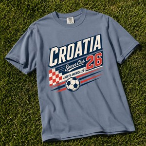 K&ouml;nnte beinhalten: Ein schieferblaues Kurzarm-T-Shirt mit einem Grafikdruck, der CROATIA Soccer Club 26 NORTH AMERICA 26 liest. Das Design umfasst ein rot-wei&szlig; kariertes Quadrat, einen Fu&szlig;ball sowie rote und blaue Bewegungslinien auf der Vorderseite.