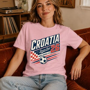 K&ouml;nnte beinhalten: Ein hellrosa T-Shirt mit kurzen &Auml;rmeln und einem Grafikdruck mit der Aufschrift CROATIA Soccer Club 26 North America 26. Das Design enth&auml;lt ein rot-wei&szlig; kariertes Quadrat, einen Fu&szlig;ball und blaue sowie rote horizontale Linien.